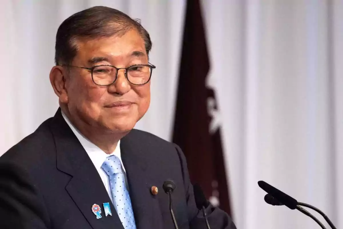 Hombre de mediana edad con gafas y traje oscuro hablando frente a un micrófono con una expresión sonriente Hombre de mediana edad con gafas y traje oscuro hablando frente a un micrófono con una expresión sonriente
