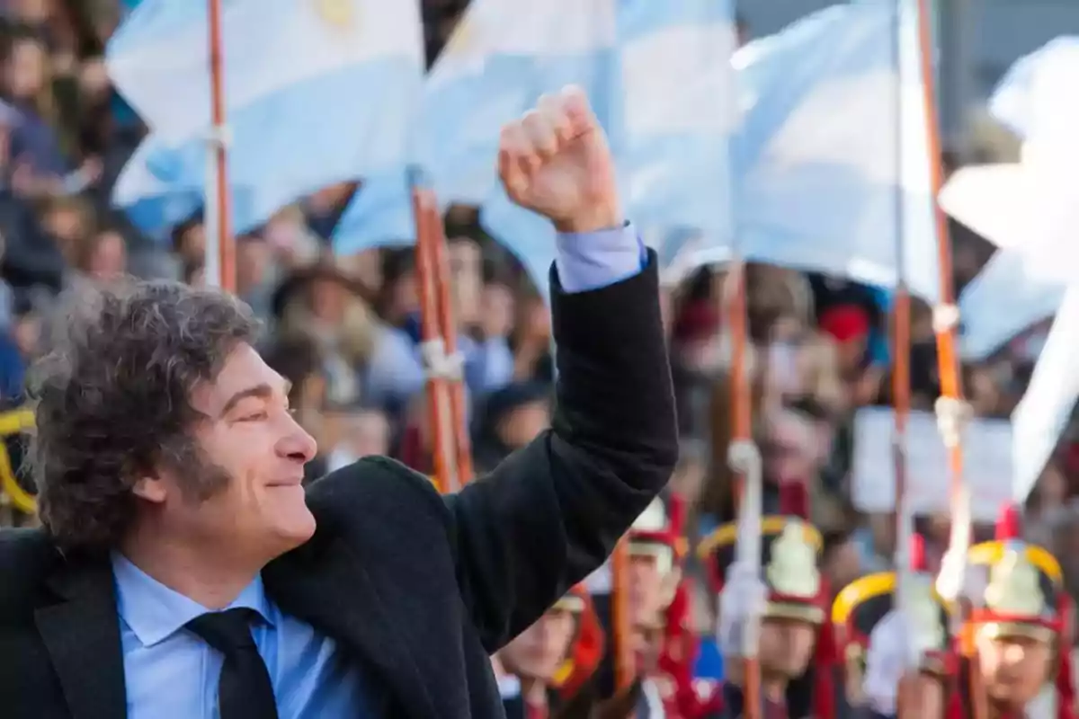 Hombre de cabello rizado con traje oscuro levantando el puño en alto frente a un grupo de personas y banderas argentinas ondeando al fondo Hombre de cabello rizado con traje oscuro levantando el puño en alto frente a un grupo de personas y banderas argentinas ondeando al fondo