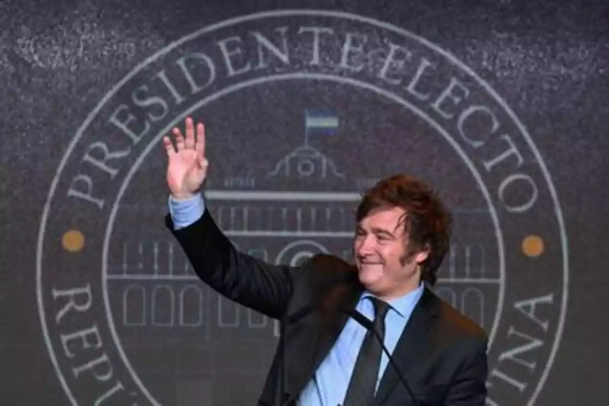 Hombre con traje saludando con la mano levantada frente a un fondo que dice presidente electo