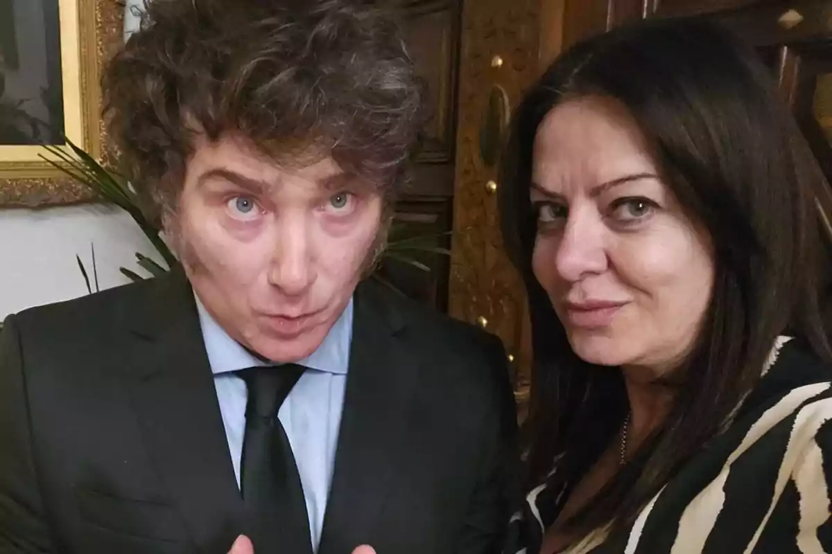 Two people pose for a selfie in an elegant indoor setting; one is wearing a suit and tie, and the other is dressed in a black and white patterned blouse.