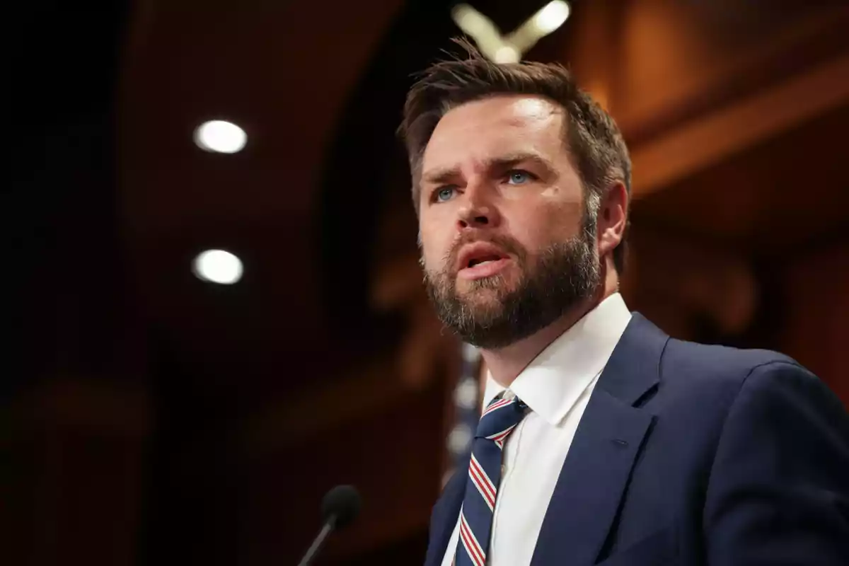 Bearded man in a blue suit speaking in front of a microphone in a formal setting