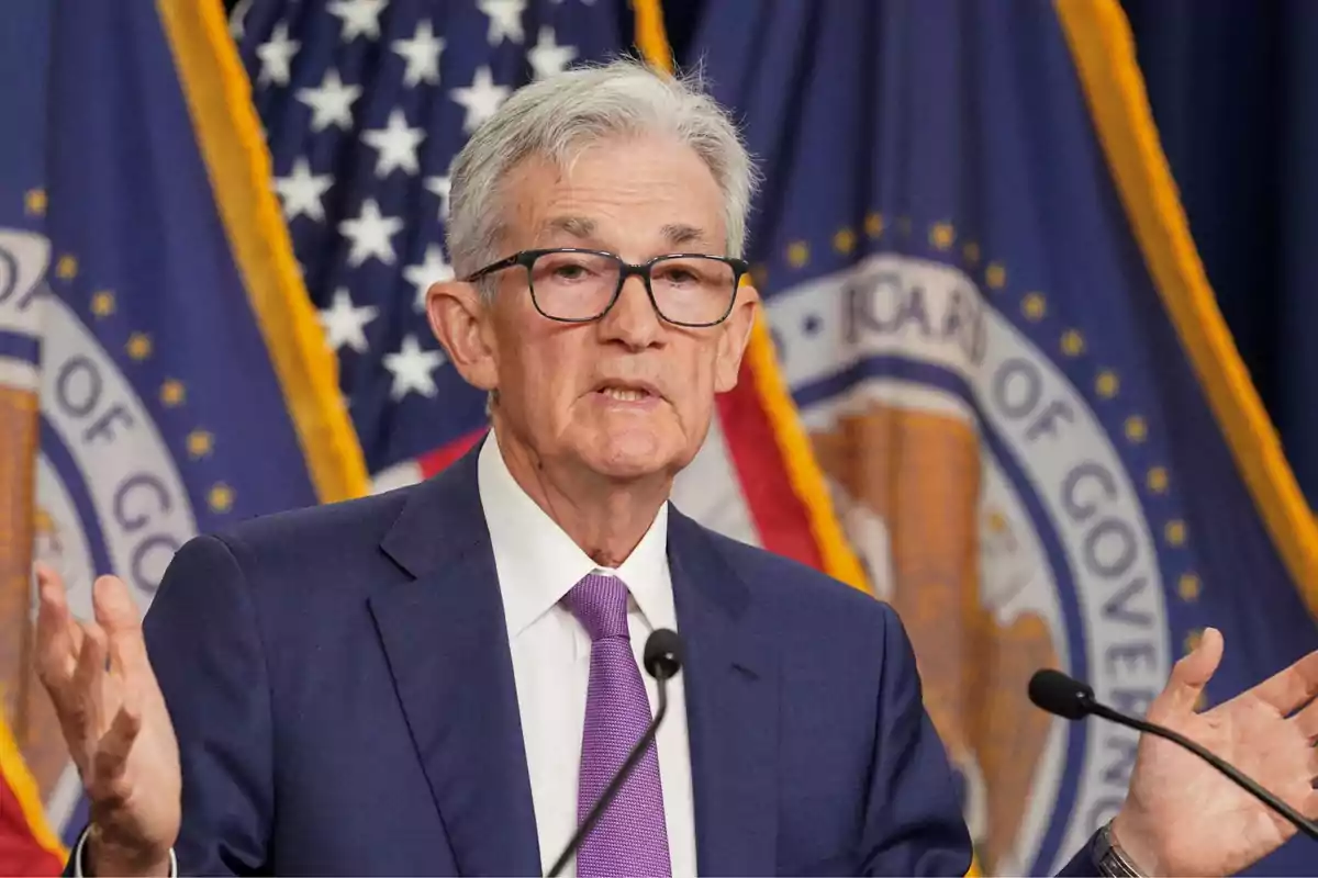 Gray-haired man with glasses speaking in front of microphones with official flags in the background