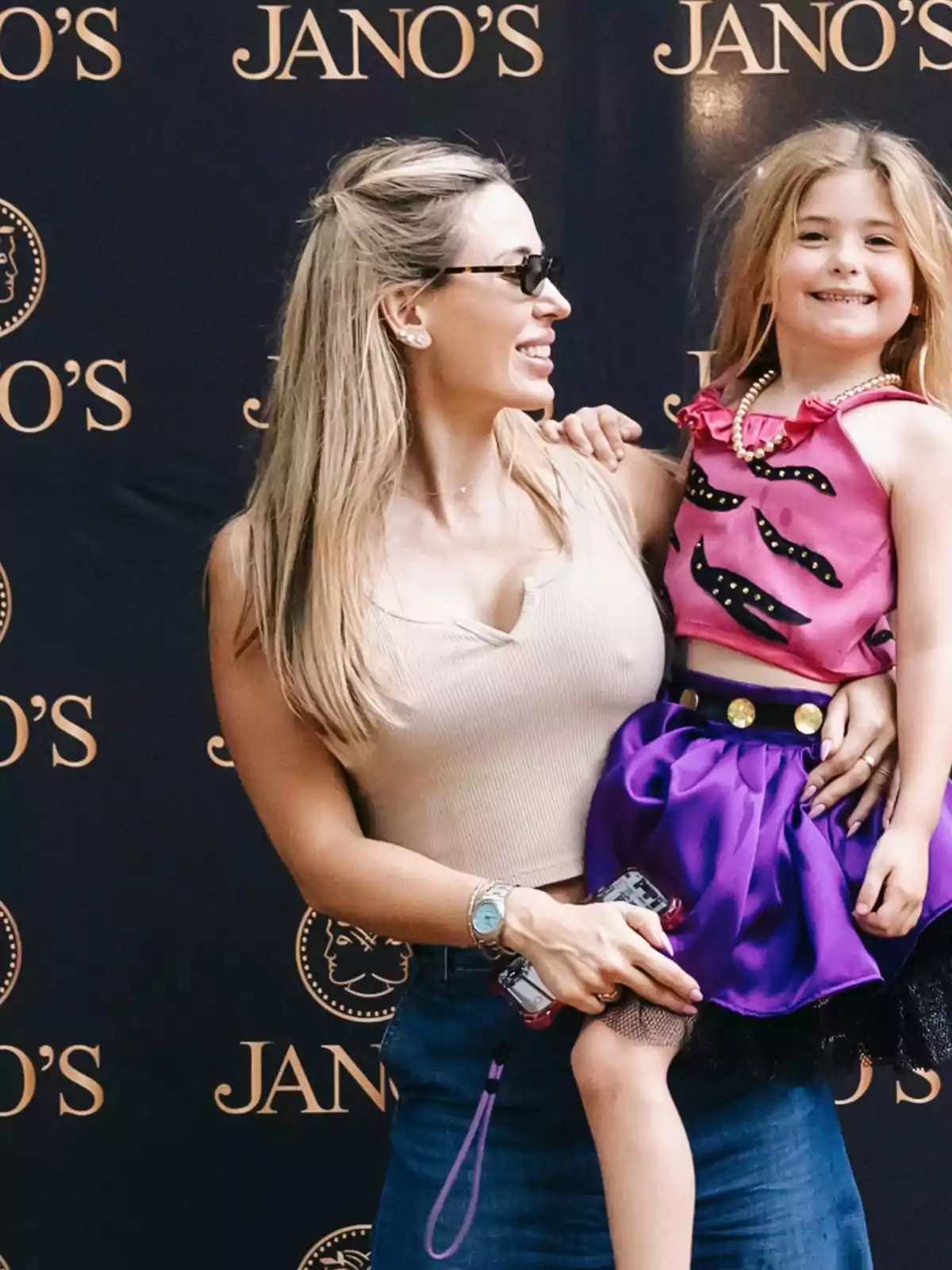 Mujer sonriente con gafas sostiene a una niña vestida con falda morada y blusa rosa frente a un fondo con el logo de Jano's Mujer sonriente con gafas sostiene a una niña vestida con falda morada y blusa rosa frente a un fondo con el logo de Jano's