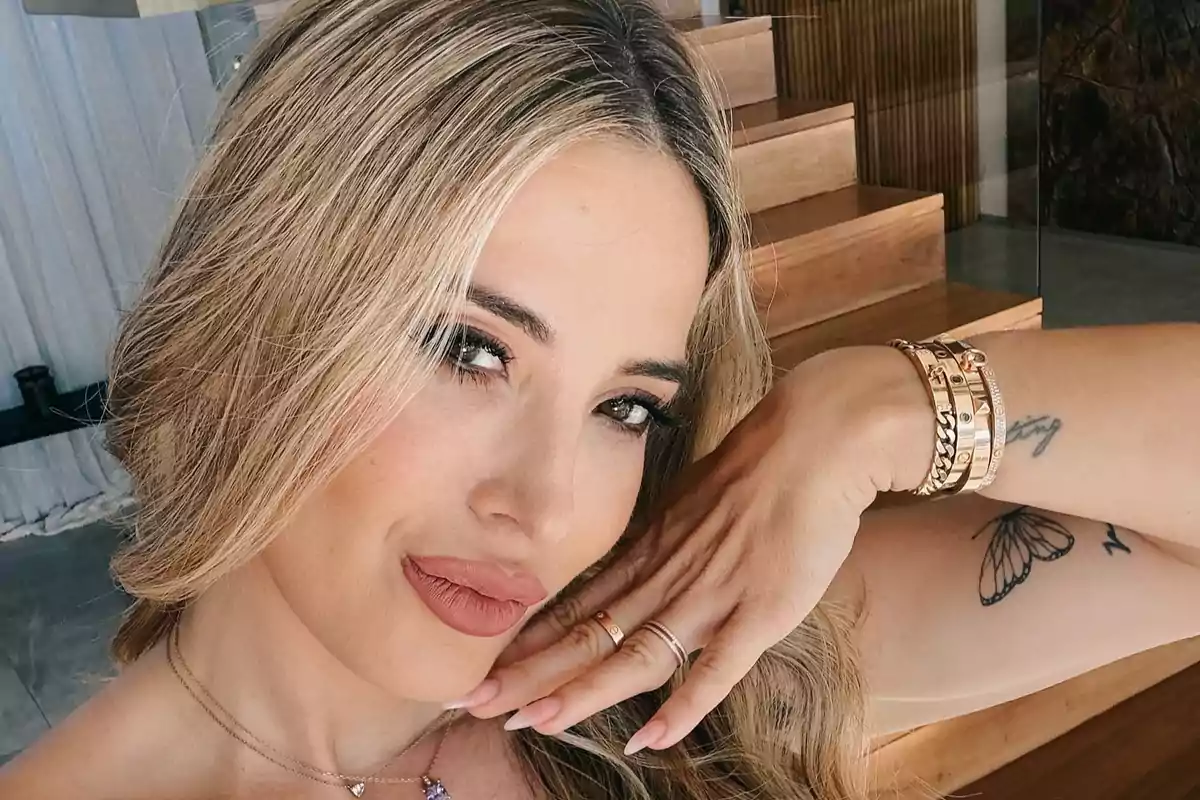Mujer rubia posando para una selfie con maquillaje natural, joyería dorada y un tatuaje de mariposa en el brazo
