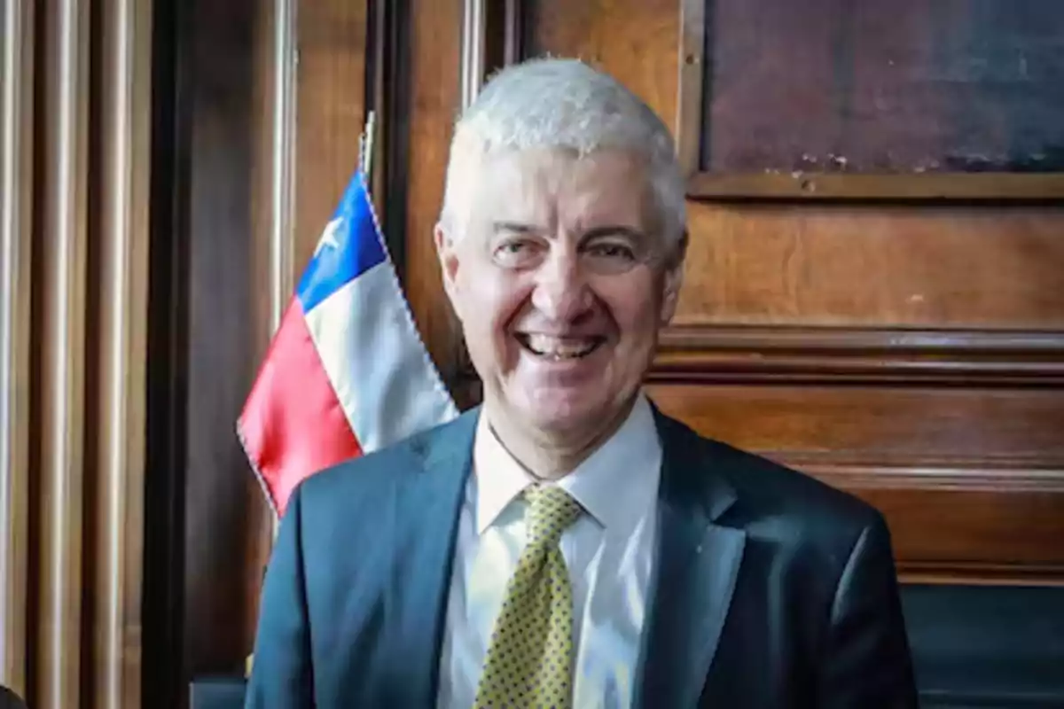 Gray-haired man in a suit and yellow tie smiling in front of a Chilean flag and a wooden background Gray-haired man in a suit and yellow tie smiling in front of a Chilean flag and a wooden background
