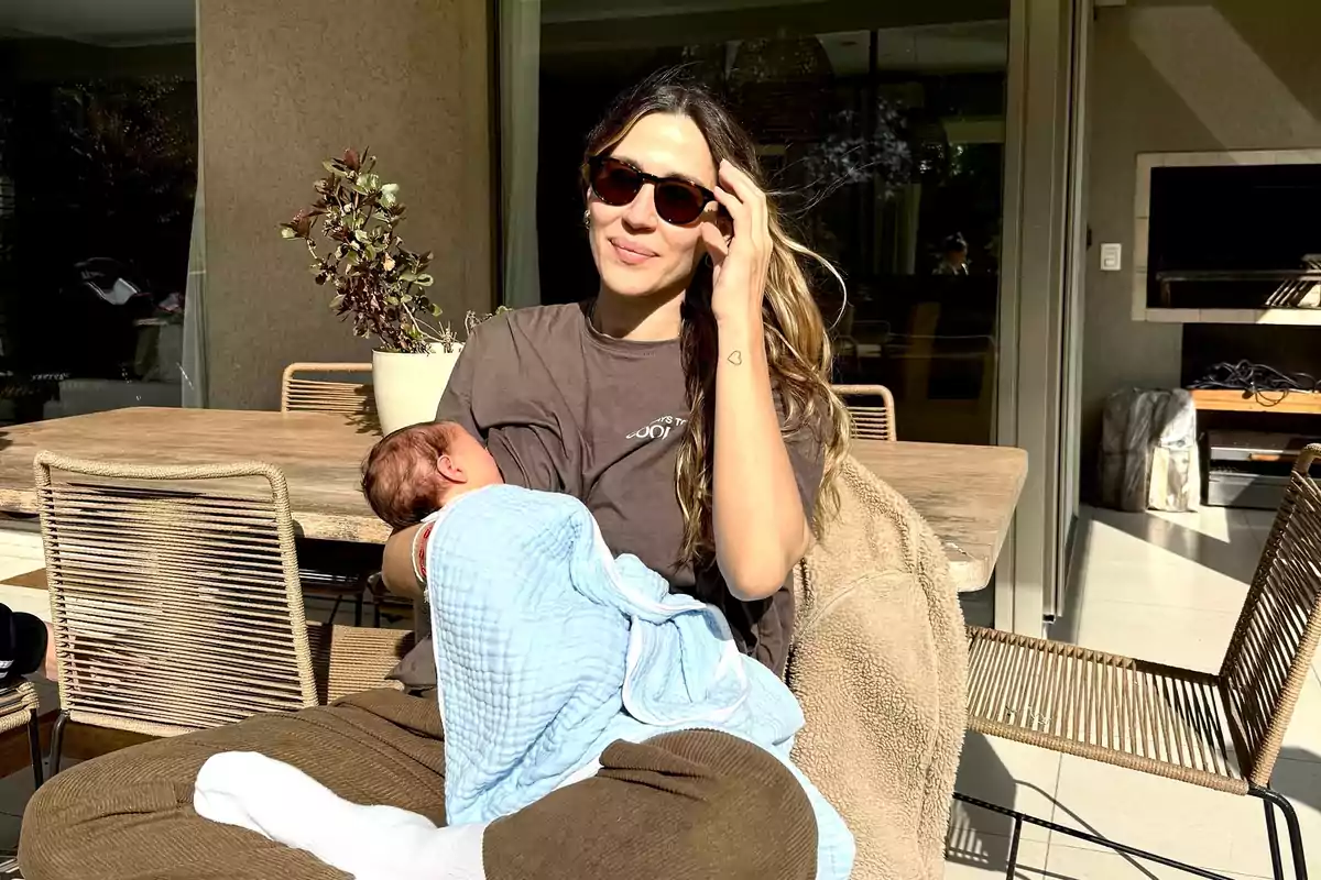 Mujer sentada al aire libre amamantando a un bebé envuelto en una manta azul mientras usa gafas de sol y sonríe
