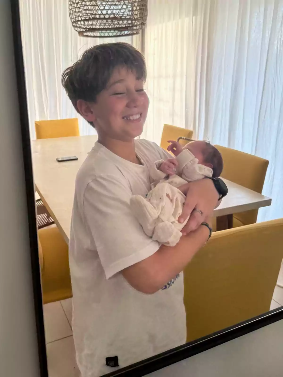 Un niño sonriente sostiene en brazos a un bebé envuelto en una manta blanca dentro de una habitación iluminada con sillas amarillas y una mesa al fondo. Un niño sonriente sostiene en brazos a un bebé envuelto en una manta blanca dentro de una habitación iluminada con sillas amarillas y una mesa al fondo.