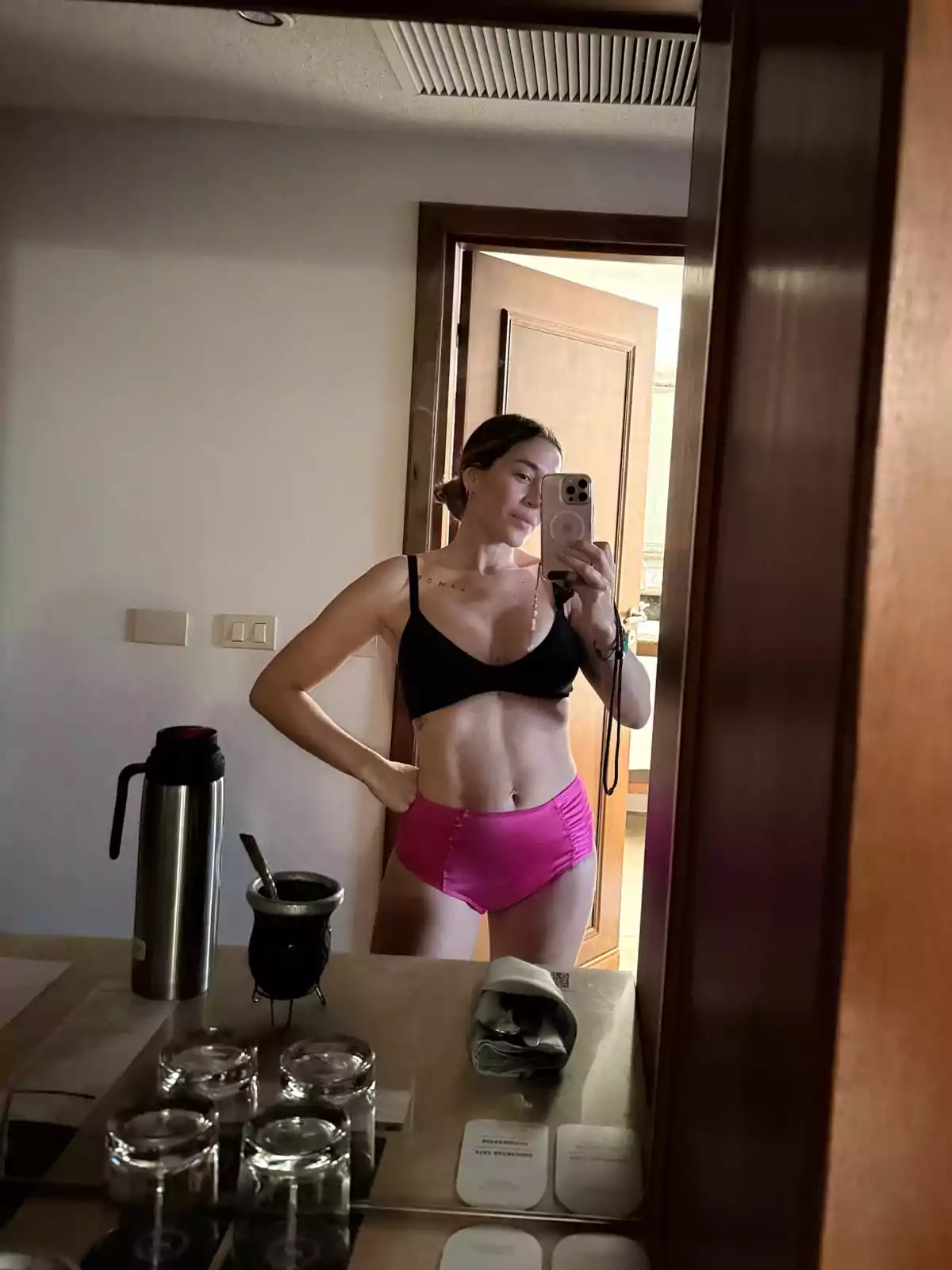 Mujer tomándose una selfie frente a un espejo usando un top negro y un short rosa en una habitación con una mesa donde hay un termo, un mate y varios vasos de vidrio