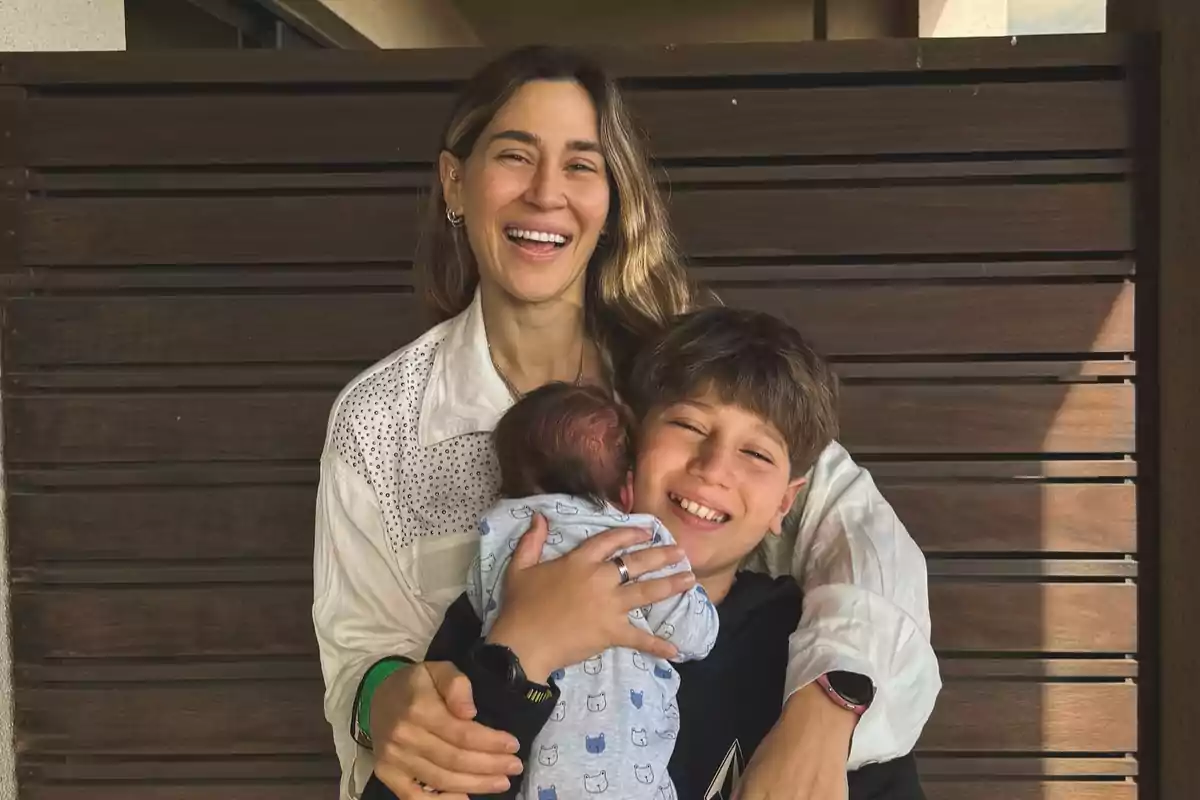 A smiling woman hugs a child and a baby in front of a wooden wall.
