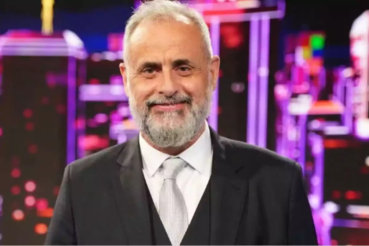 Gray-haired and gray-bearded man in a gray suit and tie smiling in front of a colorful, blurred background Gray-haired and gray-bearded man in a gray suit and tie smiling in front of a colorful, blurred background