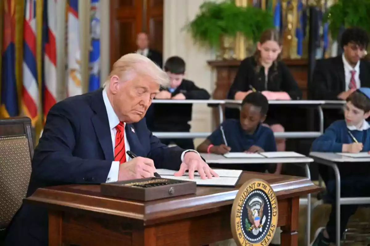 Un hombre de traje firmando un documento en un escritorio presidencial mientras varios niños escriben en pupitres detrás de él Un hombre de traje firmando un documento en un escritorio presidencial mientras varios niños escriben en pupitres detrás de él
