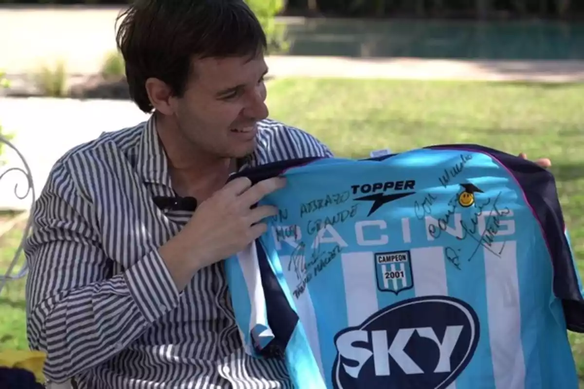 Hombre sonriente sostiene una camiseta de Racing Club firmada por varios jugadores en un entorno al aire libre Hombre sonriente sostiene una camiseta de Racing Club firmada por varios jugadores en un entorno al aire libre