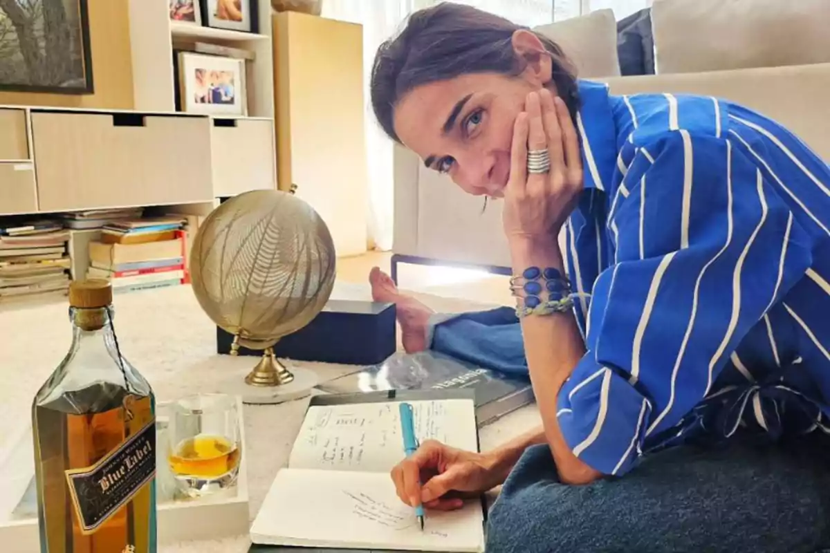 Mujer sentada en el suelo escribiendo en una libreta con una botella de whisky, un vaso, un globo terráqueo y libros a su alrededor en una sala acogedora
