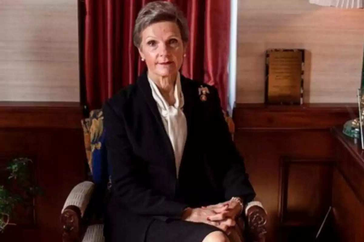 Mujer mayor de cabello corto y canoso sentada en un sillón elegante con las manos cruzadas, vestida de manera formal en un ambiente clásico con cortinas rojas y muebles de madera