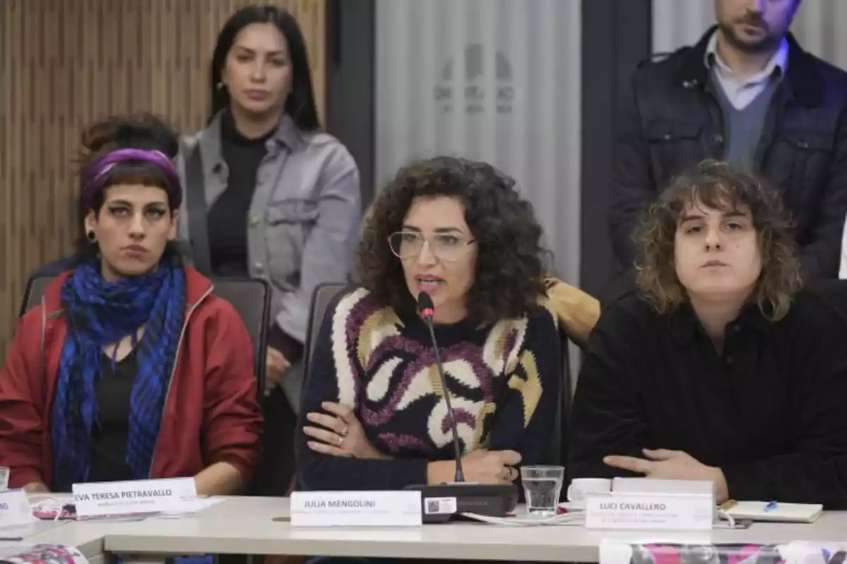Three people sitting at a table during a conference or meeting, with microphones and nameplates, while others watch from the background.