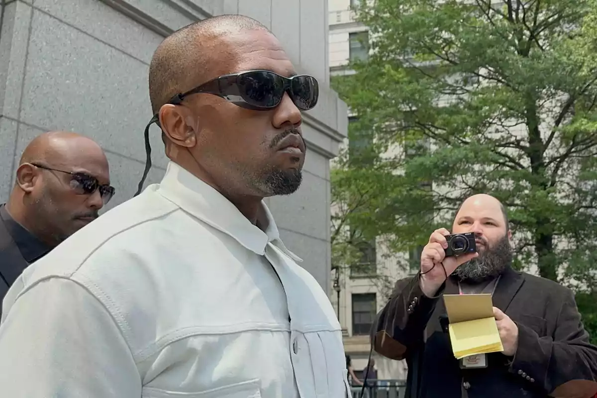 Man with sunglasses and a white jacket walking next to a security guard while a journalist photographs him and takes notes on the street in front of a building and trees