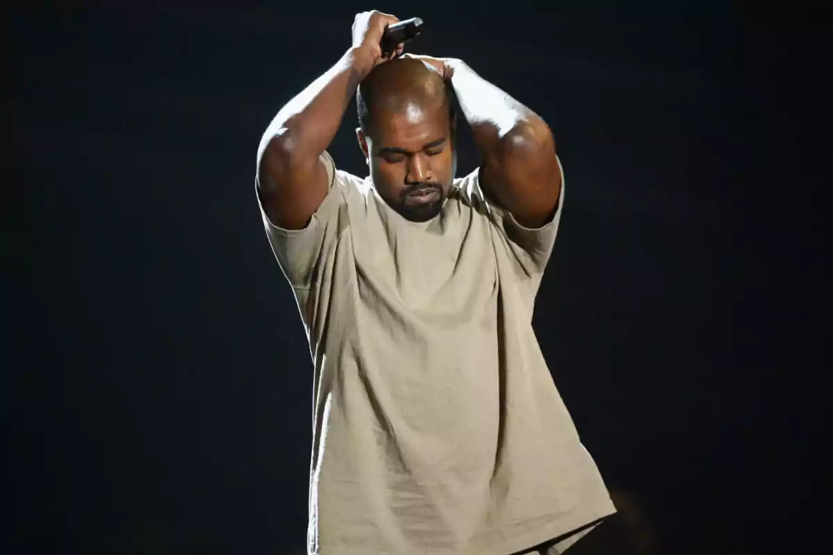 Man in a beige t-jersey holding a microphone above his head on a dark stage