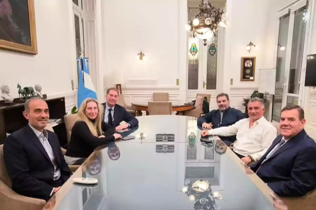 Seis personas sentadas alrededor de una mesa de reuniones en una sala elegante con una bandera argentina al fondo Seis personas sentadas alrededor de una mesa de reuniones en una sala elegante con una bandera argentina al fondo
