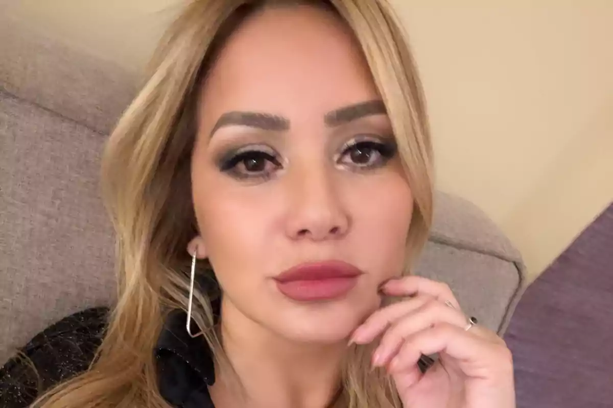Blonde woman with striking makeup and painted lips, sitting on a sofa and looking at the camera while resting her hand on her face