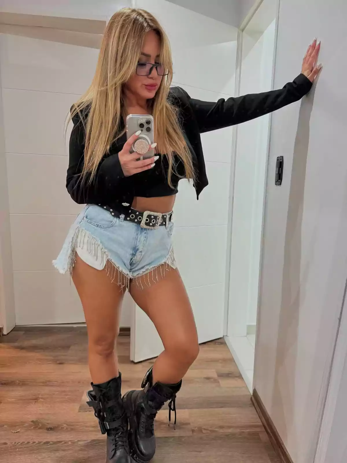 Blonde woman with glasses taking a selfie in front of a mirror, wearing a black jacket, black top, fringed denim shorts, and black boots Blonde woman with glasses taking a selfie in front of a mirror, wearing a black jacket, black top, fringed denim shorts, and black boots