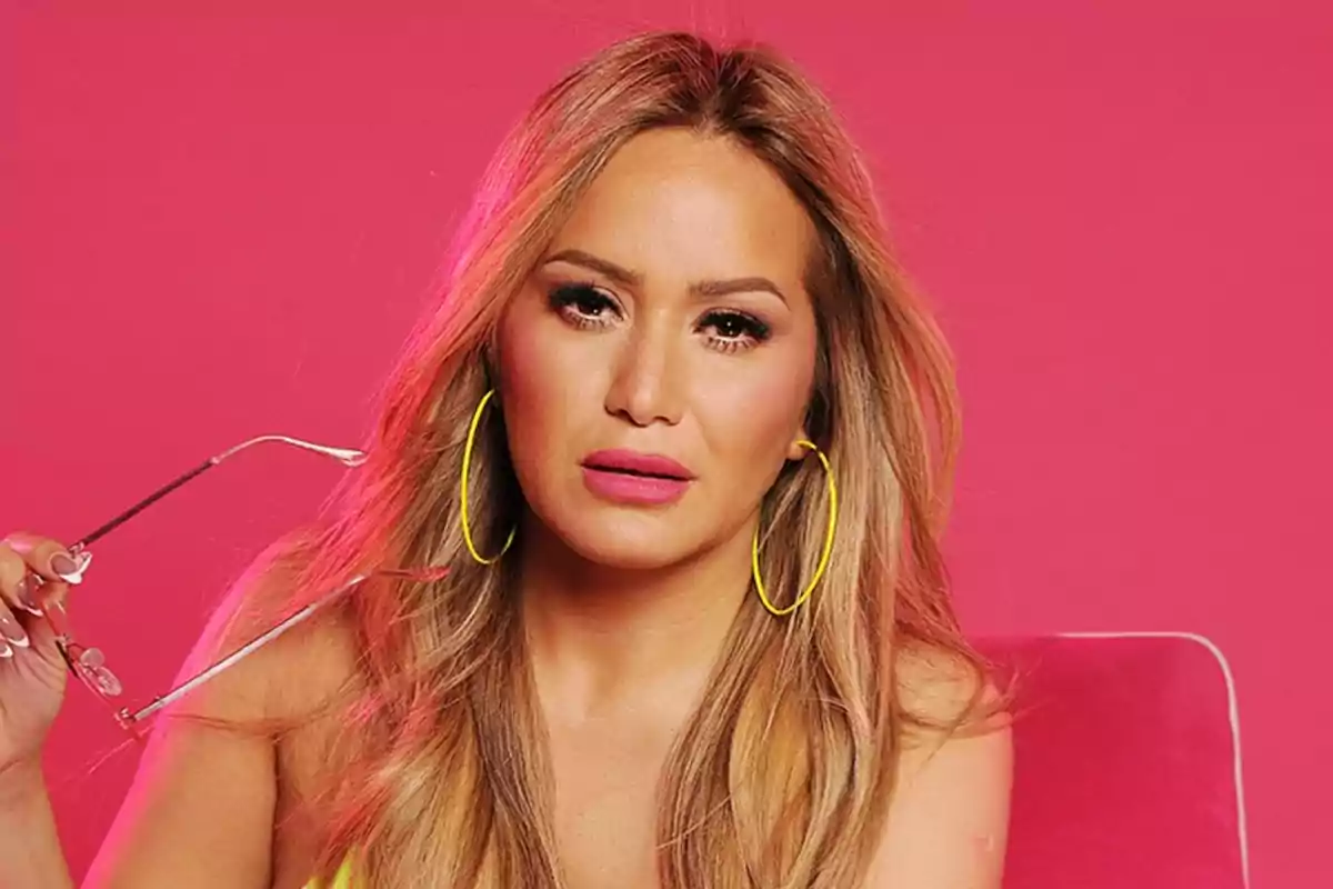 Long-haired blonde woman with large yellow earrings and pink lips holding glasses in front of a pink background