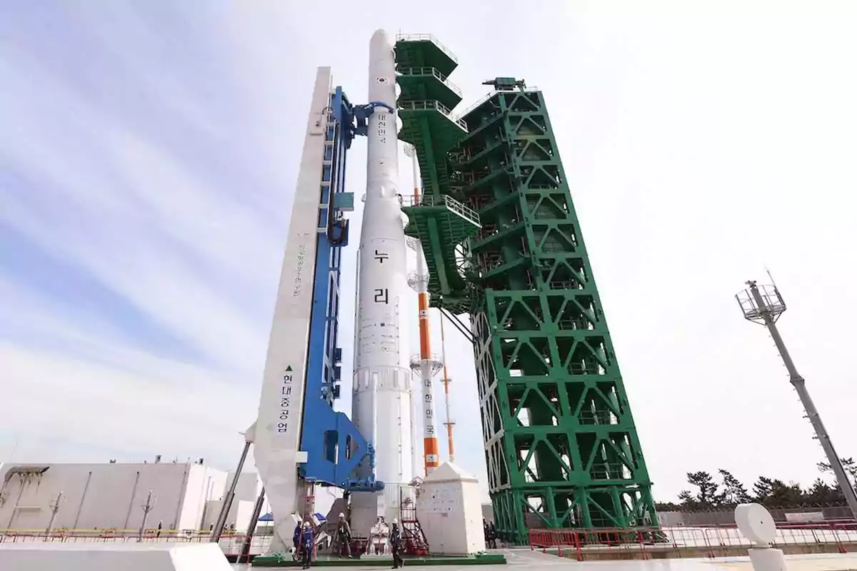 Cohete espacial blanco en posición vertical sobre una plataforma de lanzamiento con estructuras metálicas verdes y azules a su alrededor bajo un cielo parcialmente nublado Cohete espacial blanco en posición vertical sobre una plataforma de lanzamiento con estructuras metálicas verdes y azules a su alrededor bajo un cielo parcialmente nublado