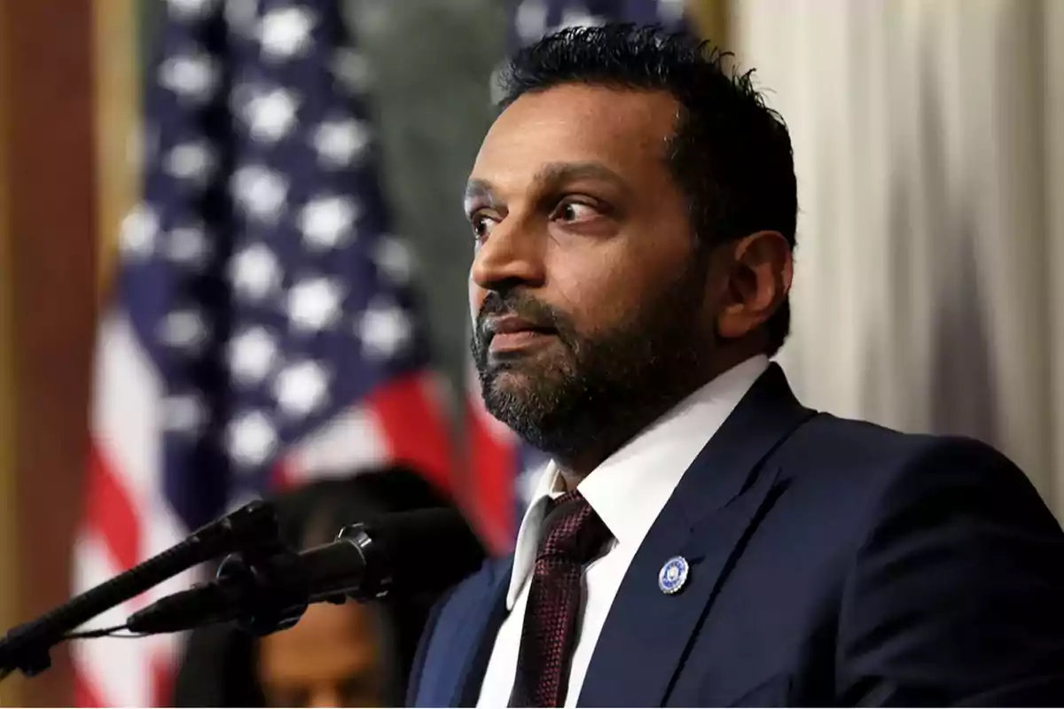 Man in a dark suit speaking in front of a microphone with a blurred United States flag in the background Man in a dark suit speaking in front of a microphone with a blurred United States flag in the background