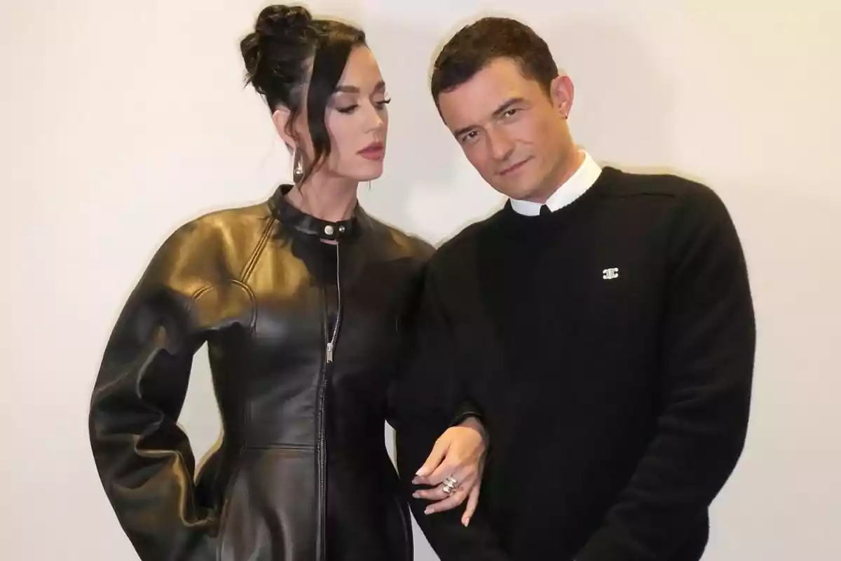 A woman in a black leather dress and a man in a black sweater pose together in front of a light-colored wall. A woman in a black leather dress and a man in a black sweater pose together in front of a light-colored wall.