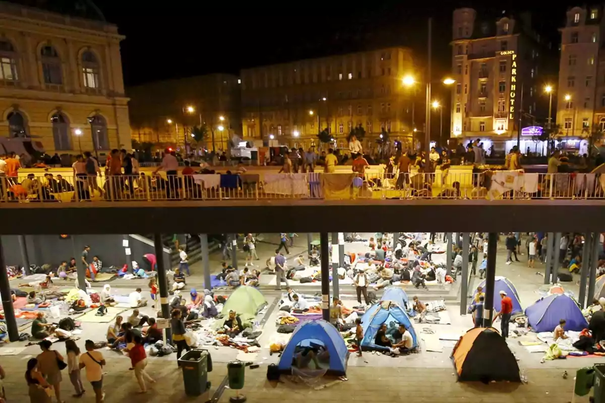 Personas acampando en una plaza urbana durante la noche rodeadas de tiendas de campaña y edificios iluminados Personas acampando en una plaza urbana durante la noche rodeadas de tiendas de campaña y edificios iluminados