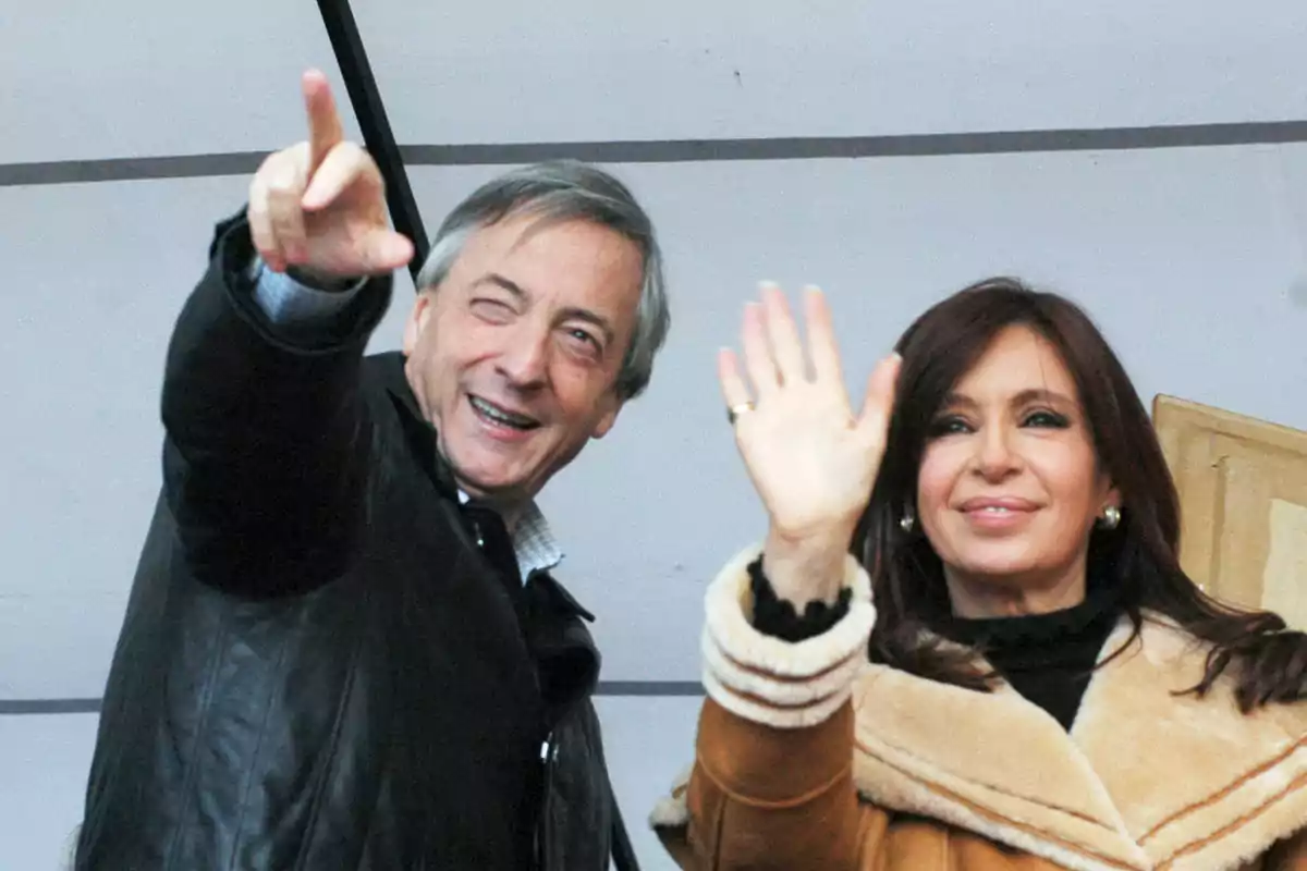 Dos personas sonríen y saludan con la mano mientras miran hacia la cámara