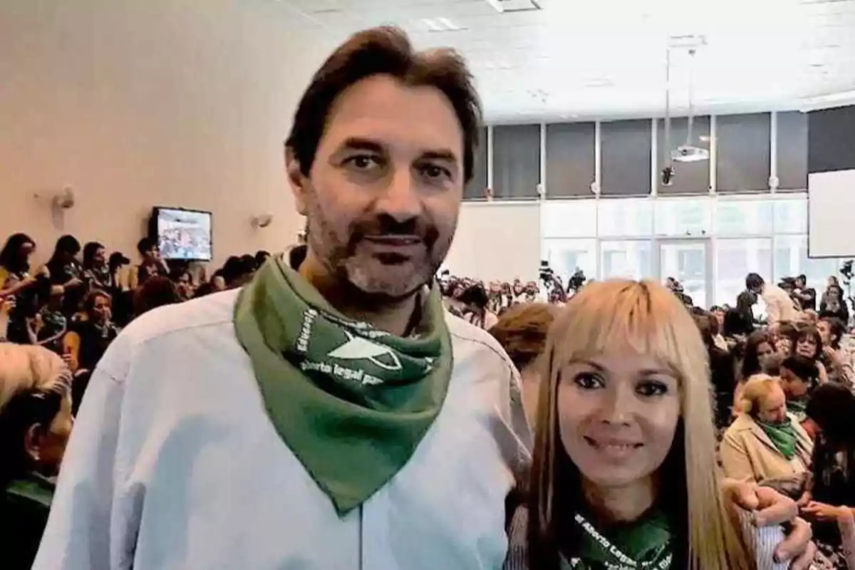 Un hombre y una mujer posan juntos en un evento con muchas personas al fondo, ambos llevan pañuelos verdes en el cuello.