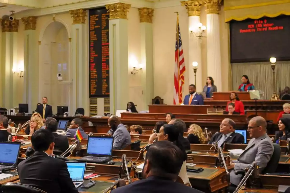 Personas reunidas en un recinto legislativo con computadoras portátiles y una bandera de Estados Unidos al fondo