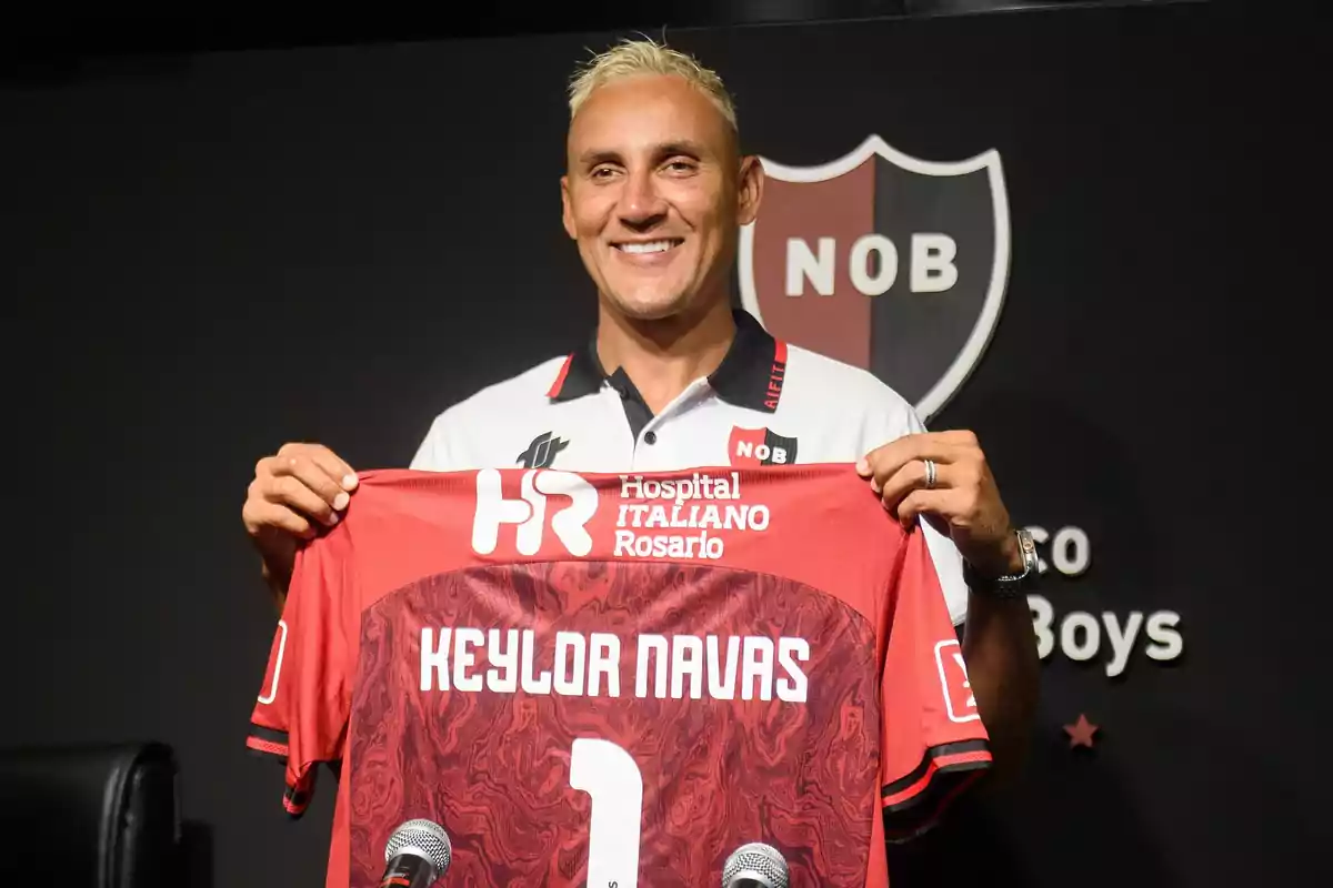 A smiling man holds a red soccer jersey with the name Keylor Navas and the number 1, in front of a background featuring the Newell's Old Boys crest. A smiling man holds a red soccer jersey with the name Keylor Navas and the number 1, in front of a background featuring the Newell's Old Boys crest.