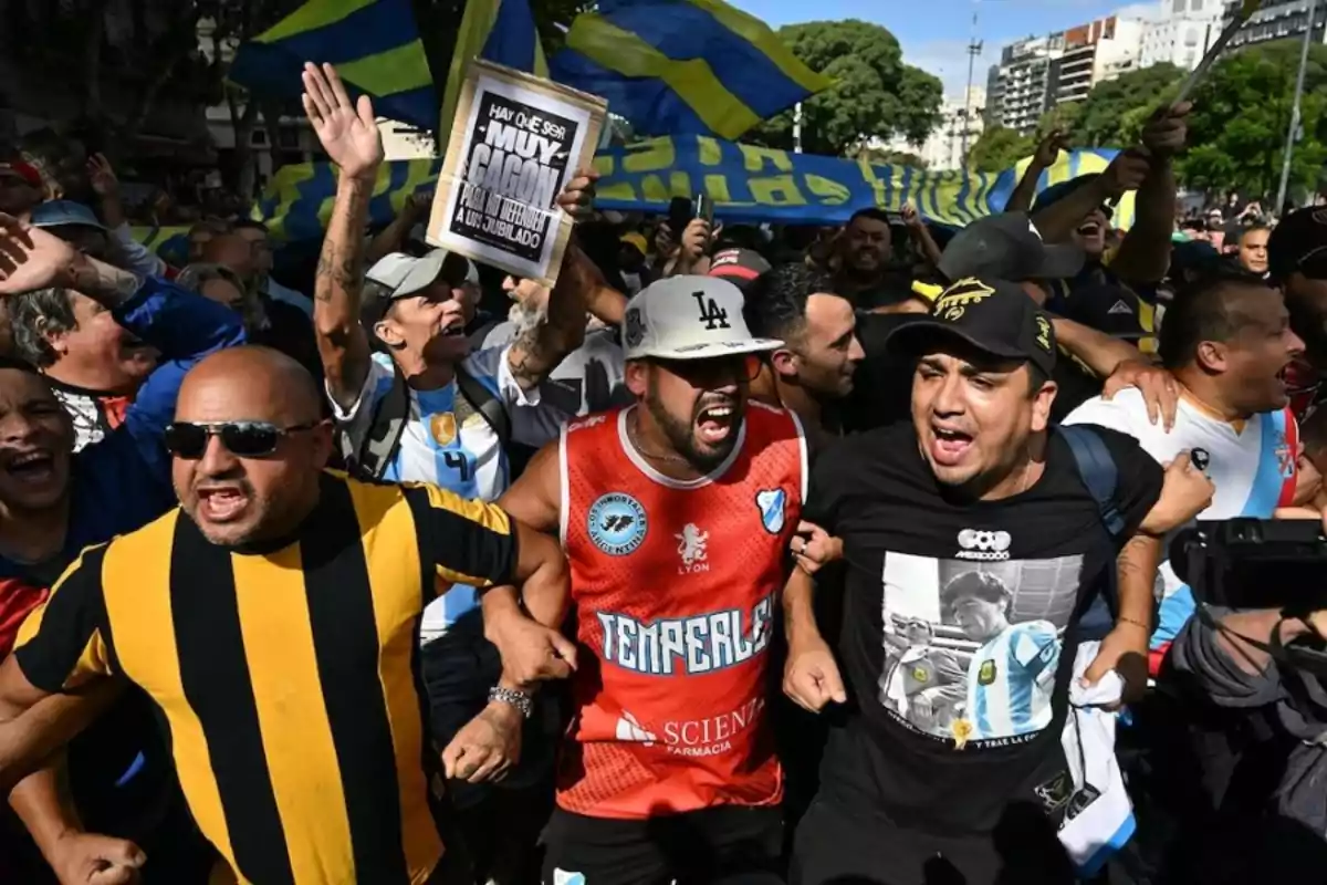 Aficionados de fútbol argentinos gritan y muestran entusiasmo durante una manifestación, algunos llevan camisetas de diferentes equipos y ondean banderas azules y amarillas en un ambiente de celebración. Aficionados de fútbol argentinos gritan y muestran entusiasmo durante una manifestación, algunos llevan camisetas de diferentes equipos y ondean banderas azules y amarillas en un ambiente de celebración.