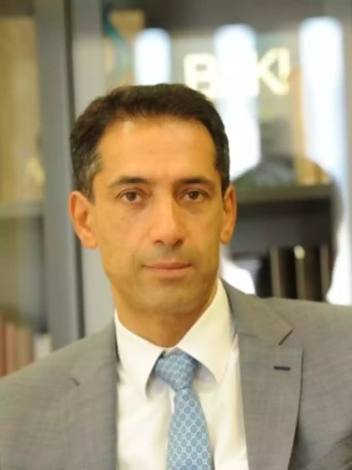 Short dark-haired man wearing a gray suit and light blue tie, posing in an office setting. Short dark-haired man wearing a gray suit and light blue tie, posing in an office setting.