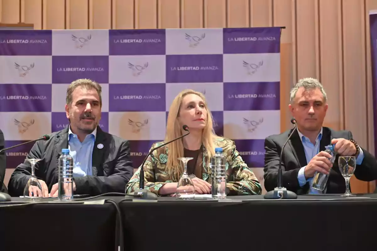 Tres personas sentadas en una mesa durante una conferencia de prensa con un fondo de logotipos de La Libertad Avanza