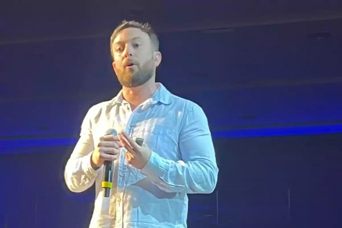 Hombre con barba y camisa clara hablando en público mientras sostiene un micrófono y un control remoto en un escenario iluminado con luces azules
