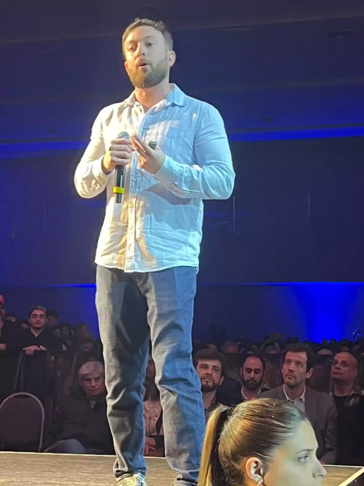 Bearded man in a light-colored jersey speaking on stage with a microphone in front of an alert audience in a setting lit with blue lights Bearded man in a light-colored jersey speaking on stage with a microphone in front of an alert audience in a setting lit with blue lights