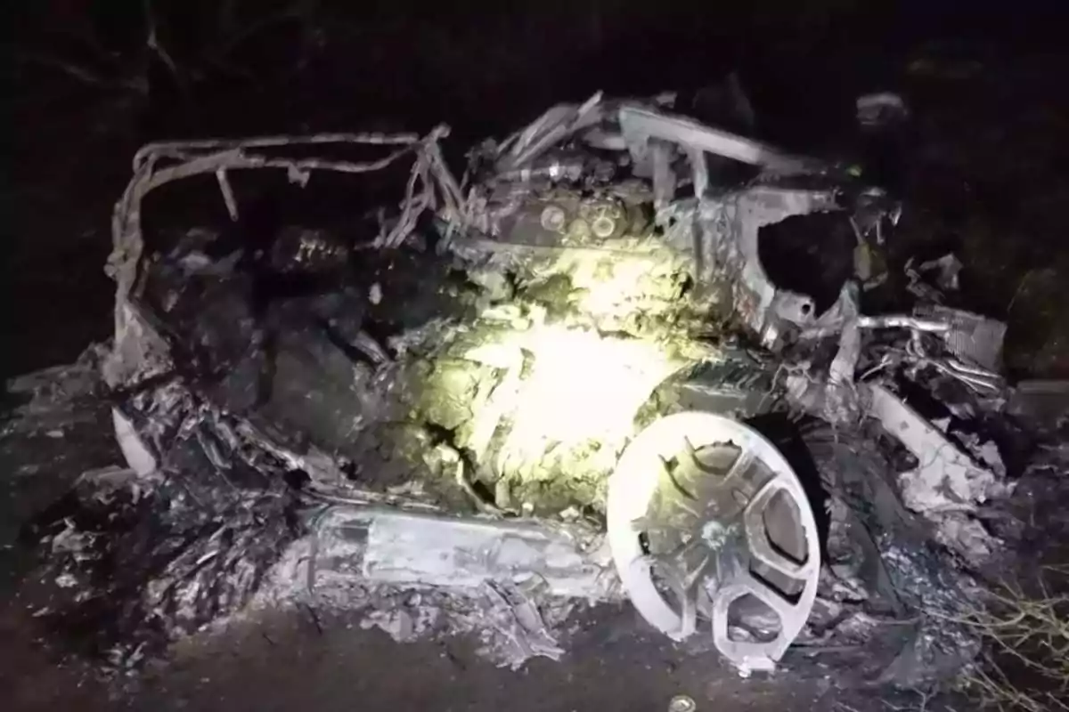 Restos calcinados de un automóvil tras un incendio en la oscuridad, con una llanta visible en primer plano y partes metálicas retorcidas Restos calcinados de un automóvil tras un incendio en la oscuridad, con una llanta visible en primer plano y partes metálicas retorcidas