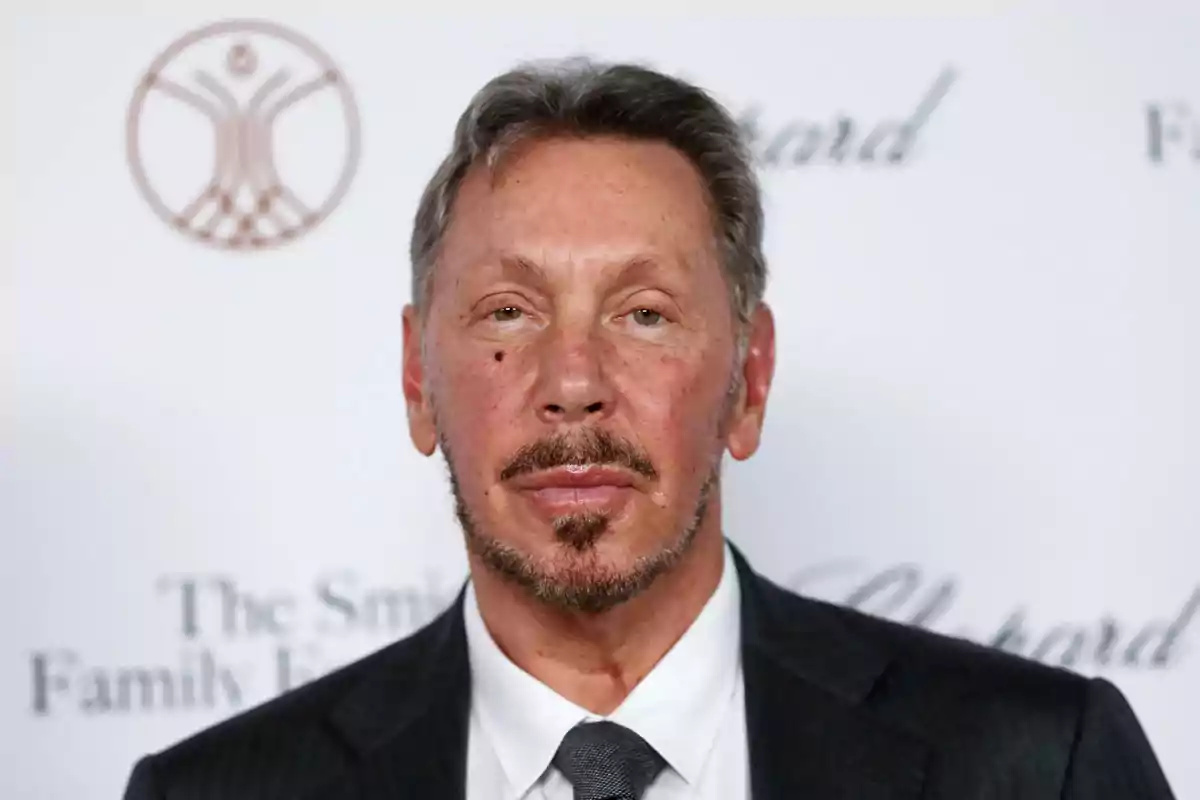 Hombre de mediana edad con barba y bigote, vestido con traje oscuro y corbata, posando frente a un fondo blanco con logotipos desenfocados. Hombre de mediana edad con barba y bigote, vestido con traje oscuro y corbata, posando frente a un fondo blanco con logotipos desenfocados.