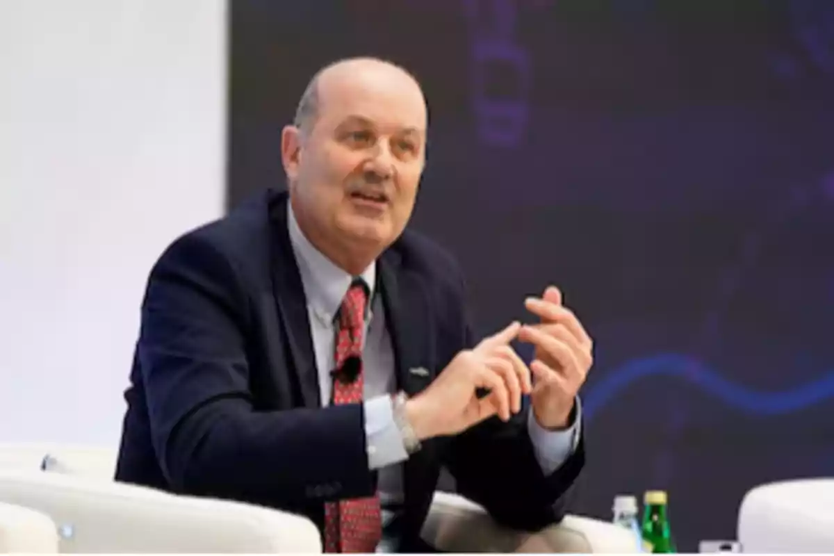 Middle-aged bald man in a dark suit and red tie speaking and gesturing while seated in a white armchair during an event or conference Middle-aged bald man in a dark suit and red tie speaking and gesturing while seated in a white armchair during an event or conference