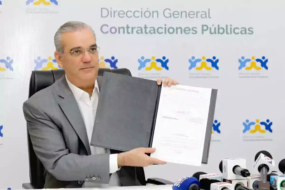 Hombre de cabello canoso y gafas sentado en una mesa mostrando un documento firmado en una carpeta negra frente a varios micrófonos con un fondo que dice Dirección General de Contrataciones Públicas Hombre de cabello canoso y gafas sentado en una mesa mostrando un documento firmado en una carpeta negra frente a varios micrófonos con un fondo que dice Dirección General de Contrataciones Públicas
