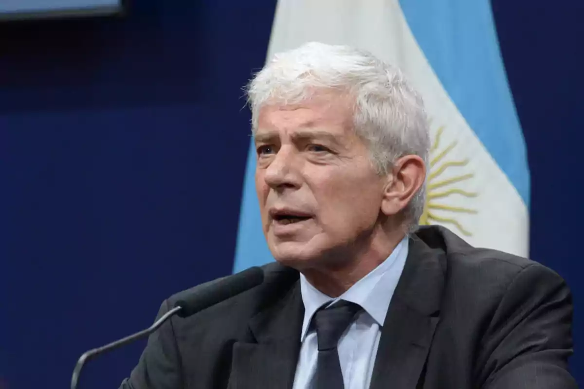 Hombre de cabello canoso hablando frente a un micrófono con una bandera de Argentina de fondo