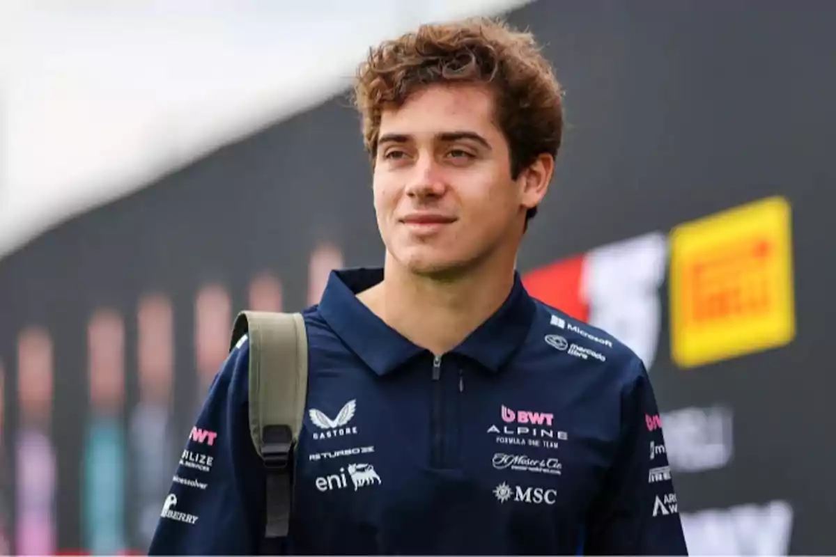 Joven con cabello rizado y camiseta azul marino con logos de patrocinadores, lleva una mochila y está en un entorno relacionado con automovilismo.