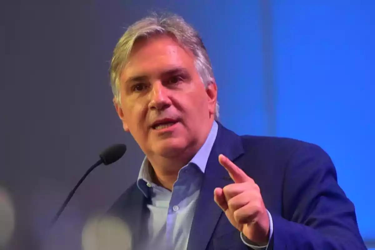Gray-haired man speaking into a microphone and pointing with his finger while wearing a blue jacket and a light-colored jersey Gray-haired man speaking into a microphone and pointing with his finger while wearing a blue jacket and a light-colored jersey