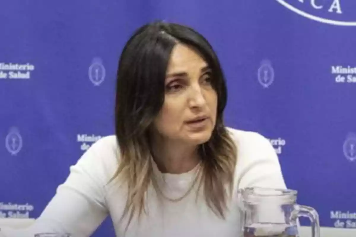 Mujer de cabello oscuro y lacio sentada frente a un fondo azul con el texto Ministerio de Salud y un vaso de vidrio sobre la mesa