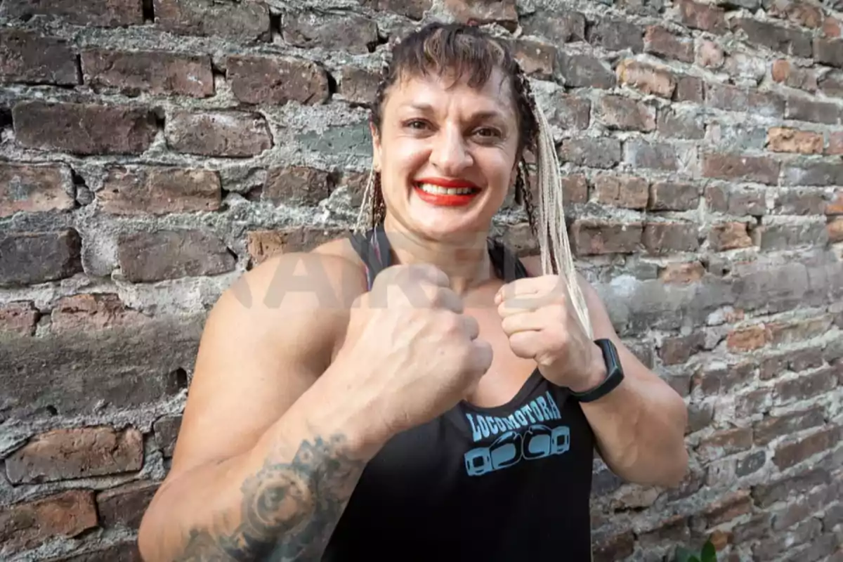 Mujer musculosa con camiseta negra y trenzas rubias sonríe mostrando los puños frente a una pared de ladrillos