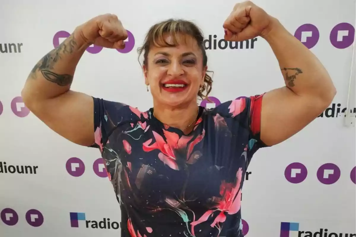 Mujer sonriente con labios rojos y camiseta de flores mostrando sus músculos frente a un fondo con logotipos de radiounr Mujer sonriente con labios rojos y camiseta de flores mostrando sus músculos frente a un fondo con logotipos de radiounr
