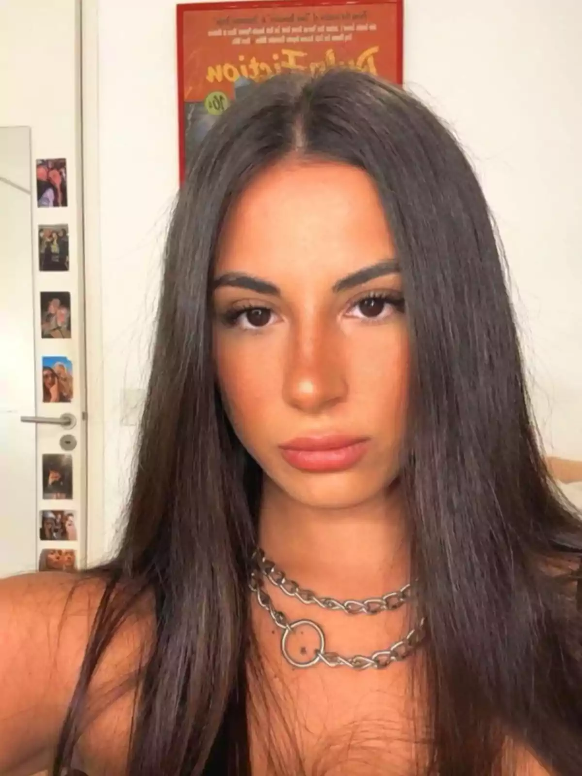Young person with long, straight hair and a silver chain necklace posing in front of the camera in a room with photos on the door and a poster on the wall Young person with long, straight hair and a silver chain necklace posing in front of the camera in a room with photos on the door and a poster on the wall