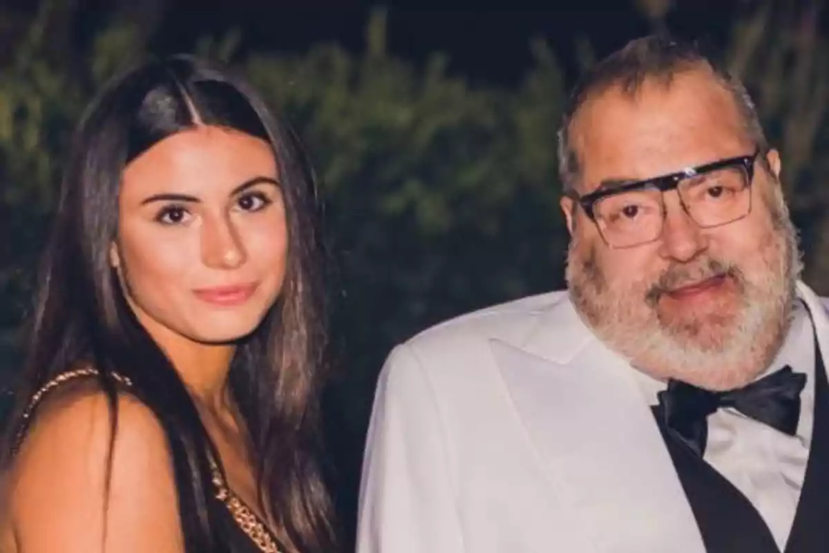 A young woman with long dark hair poses next to an older man with a beard and glasses who is wearing a white tuxedo. A young woman with long dark hair poses next to an older man with a beard and glasses who is wearing a white tuxedo.